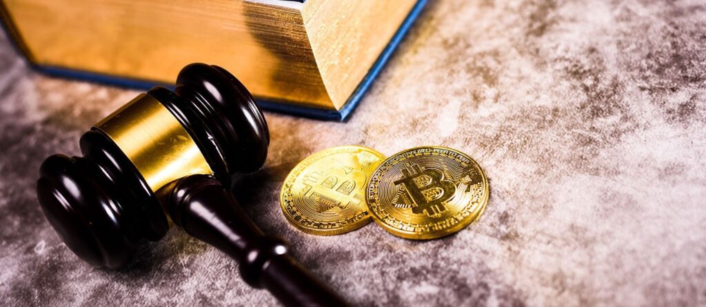 An image containing a gavel, two coins with inscriptions of bitcoin and a book on a desk.
