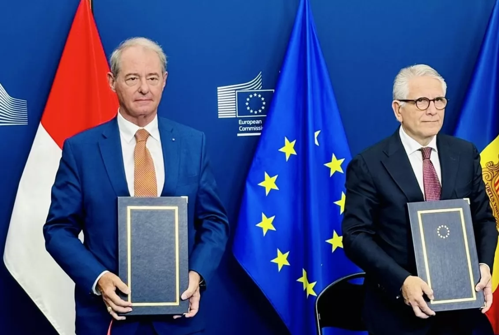 Two officials holding up agreements in front of an EU and Monaco flag