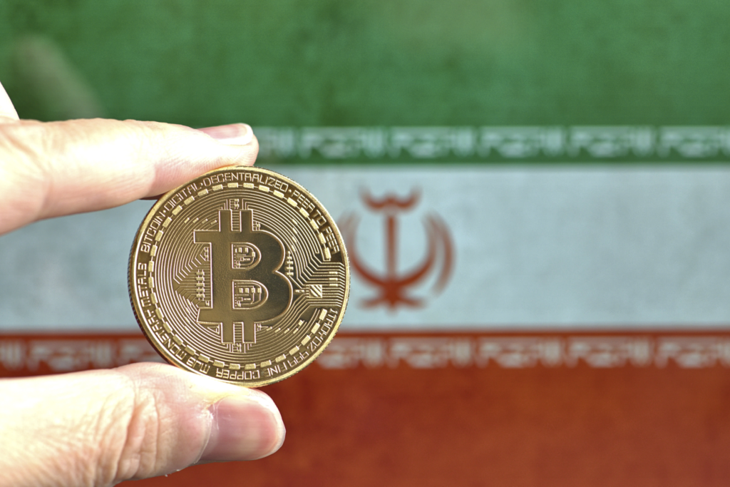 Two fingers holding up a golden bitcoin against an Iranian flag in the background