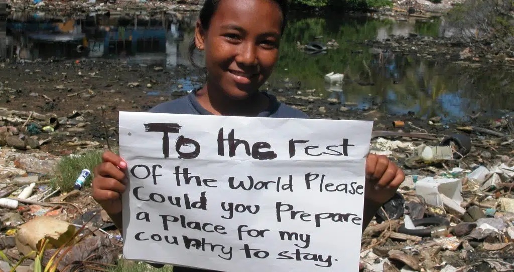 A little girl from Tuvalu holding a sign tagged "To the rest of the world please could you prepare a place for my country to stay".