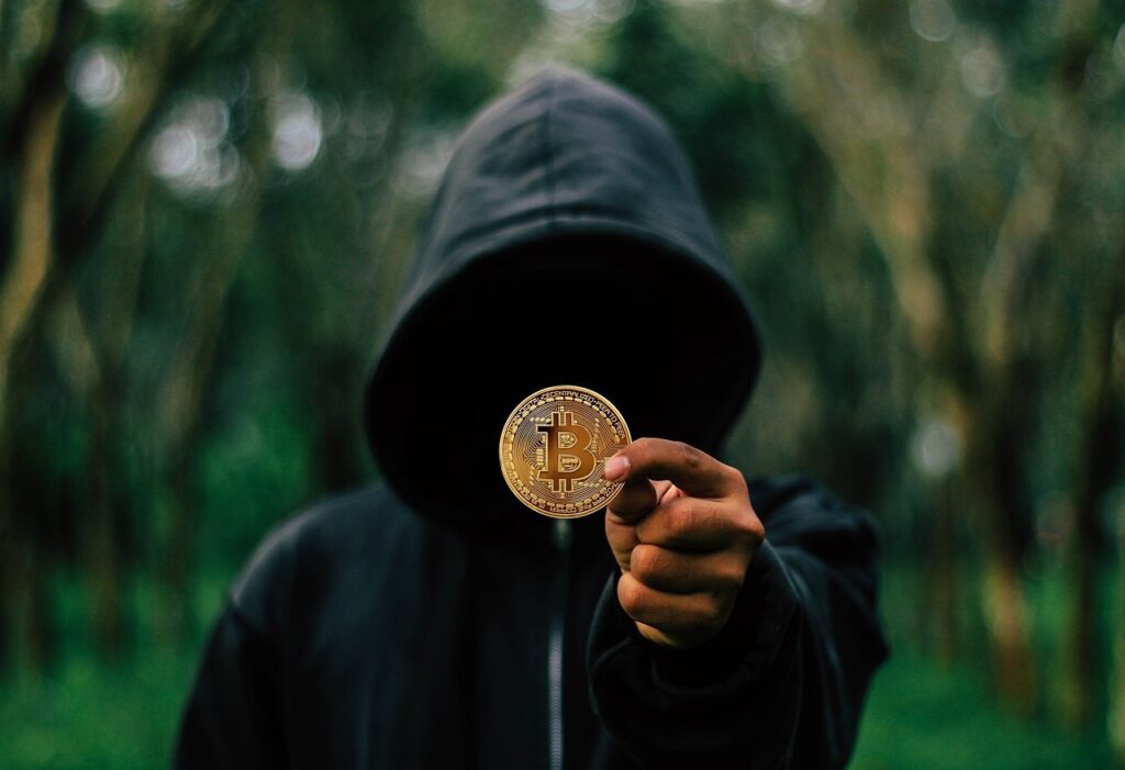A person holding a single golden Bitcoin coin in front of their face while wearing a black hoodie and standing in a blurry green forest