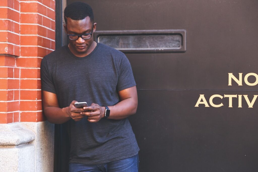 A black man in glasses wearing a casual outfit and leaning next to a wall. His eyes are glued to his phone. 